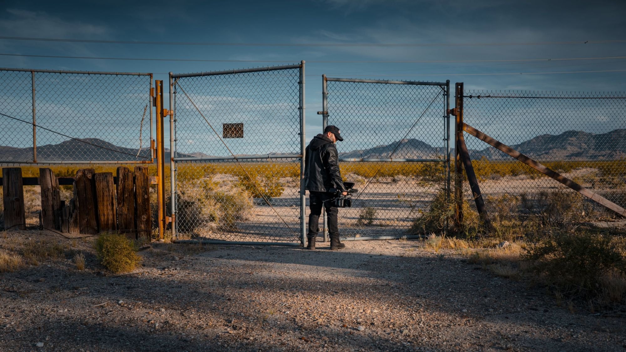 Roberto Lee Cortes holding a cinema camera at a chain-link gate in the desert at golden hour